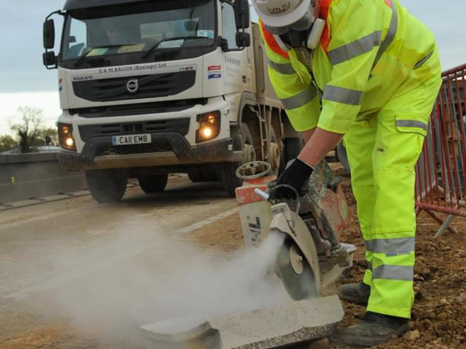 Construction worker in high-visibility yellow protective clothing and a white hard hat uses a concrete-cutting saw to trim a kerb at a roadside work site. Dust rises from the cut while a white Volvo truck with amber warning lights is parked on the road behind. Overcast sky and temporary safety barriers are visible in the background.