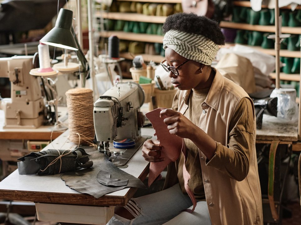 Person working at a sewing machine in a small workshop, carefully handling a piece of material with tools and supplies visible around the workspace.