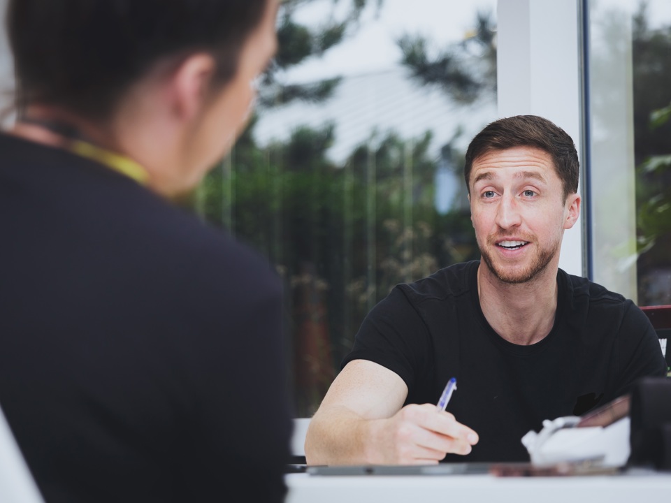A person sits at a table writing while speaking with another individual across from them in a bright, indoor setting.