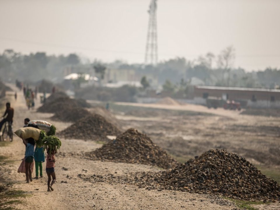 Several people walk along a wide dirt road carrying bags and bundles. Piles of gravel or stones line the roadside at regular intervals, while a cyclist travels in the same direction. The landscape appears dry and open, with scattered buildings, trees, and a communications tower visible in the hazy background.