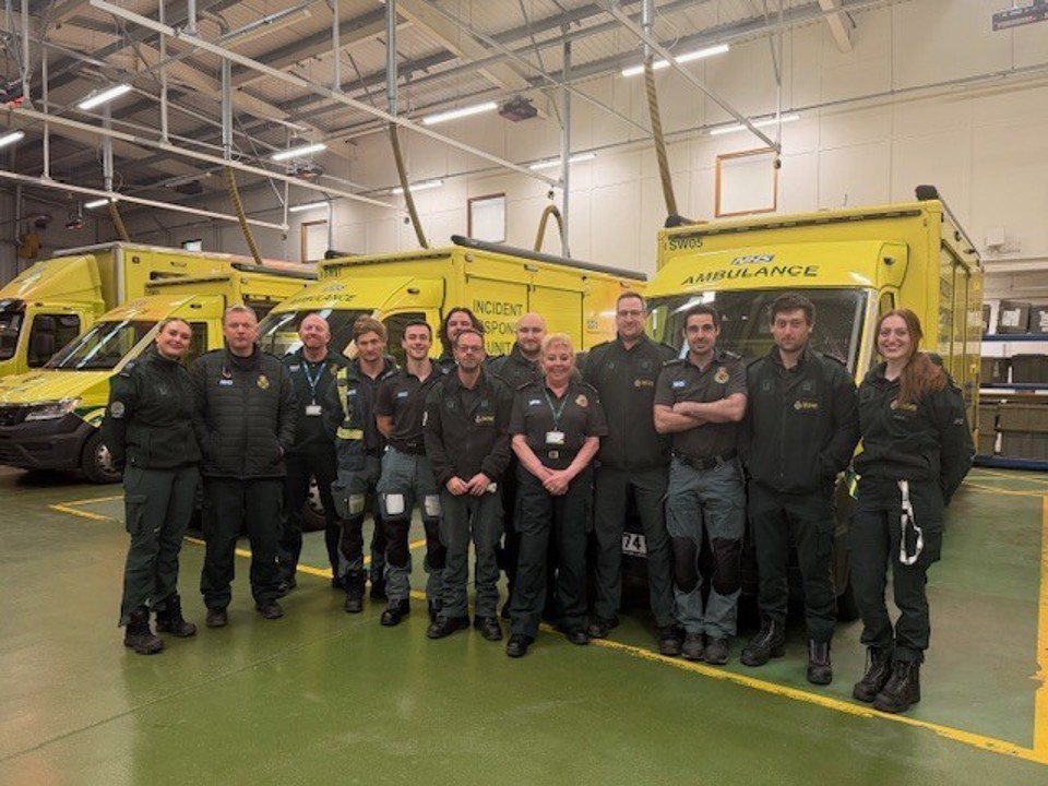 Group of ambulance service staff in uniform standing inside a vehicle workshop, positioned in front of three yellow ambulances parked side by side under overhead lighting and equipment.