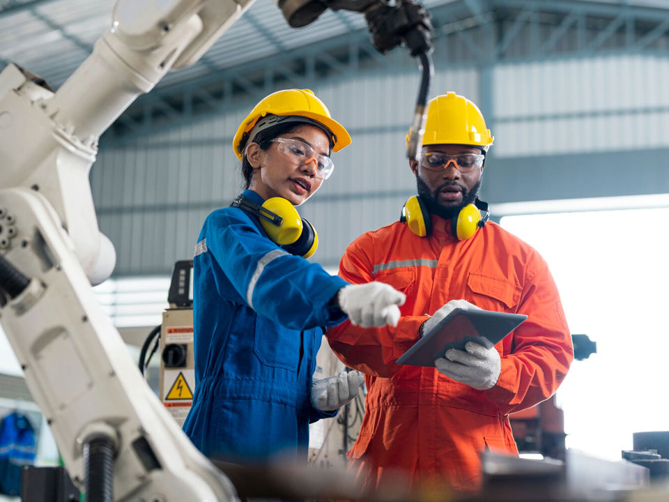 Two workers in protective gear looking at tablet next to a robot arm in high-tech factory