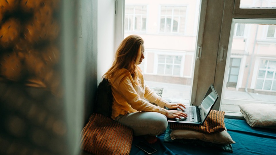 Young woman sitting on bed by window working at laptop
