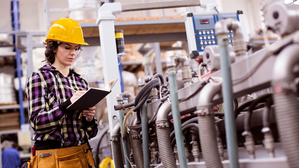 Woman in hard hat standing next to factory machine and making notes on clipboard