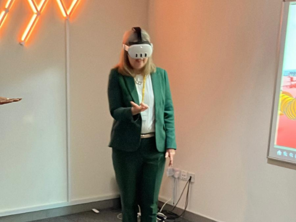 A person wearing a green suit with a white blouse stands in a modern indoor setting, holding a smartphone in one hand. The room has gray carpet flooring and light-colored walls. On the left wall, there is a decorative orange neon design resembling angled lines. To the right, a wall-mounted screen displays colorful graphics. Electrical cables and a socket are visible near the floor.