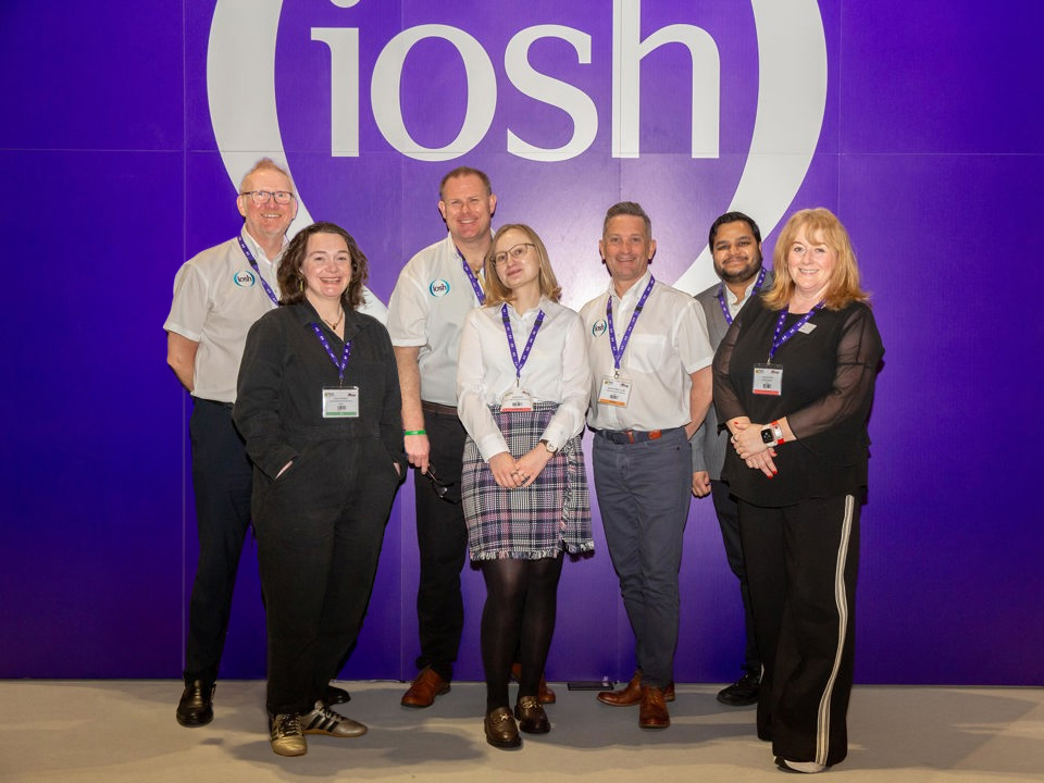 Group photo of eight conference attendees standing in front of a large purple exhibition wall displaying the white "iosh" logo. The attendees wear event lanyards and business or smart-casual clothing, with several wearing white shirts featuring the IOSH logo. The group is posed facing the camera in a well-lit indoor event space.