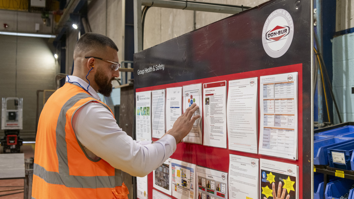 Employee wearing a high-visibility vest and ear protection reviewing health and safety information on a Don-Bur noticeboard inside the factory.