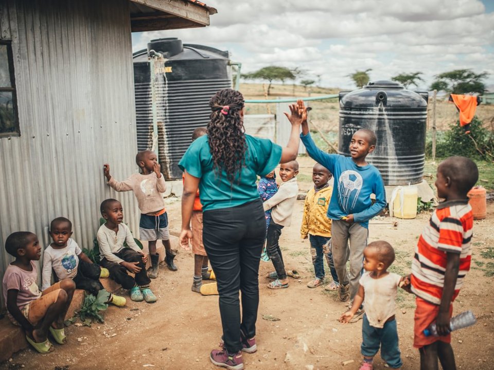 A group of children and an adult stand outside a corrugated metal building in a rural outdoor setting. The adult and one child are meeting hands in a high five while several other children watch or play nearby. Large black water storage tanks are visible behind the group, with a dry landscape, scattered trees, and cloudy skies in the background.