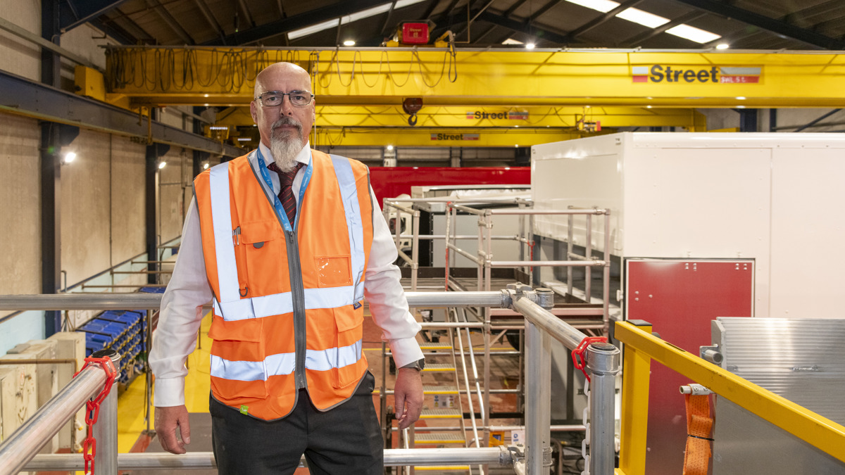 Mark Nixon standing on a factory floor at Don-Bur Bodies & Trailers, wearing a high-visibility vest and positioned in front of industrial equipment and overhead cranes.