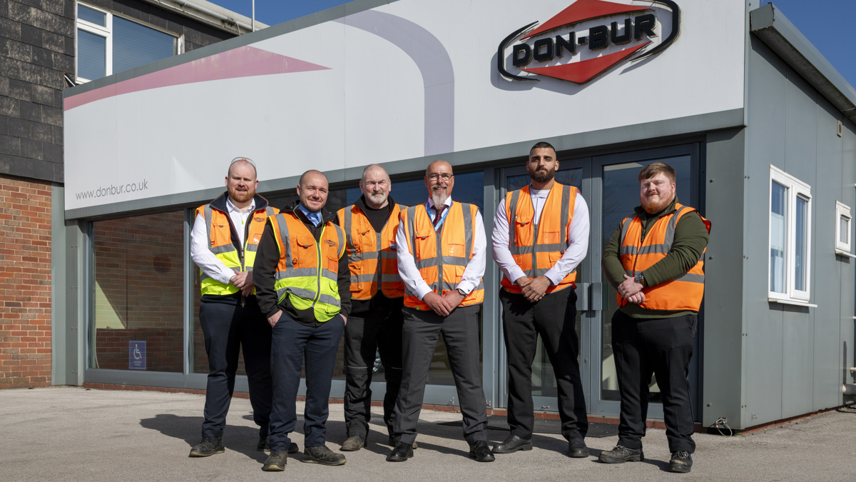 Group of six employees wearing high-visibility vests standing outside the Don-Bur Bodies & Trailers Ltd building, with the company logo displayed above the entrance.