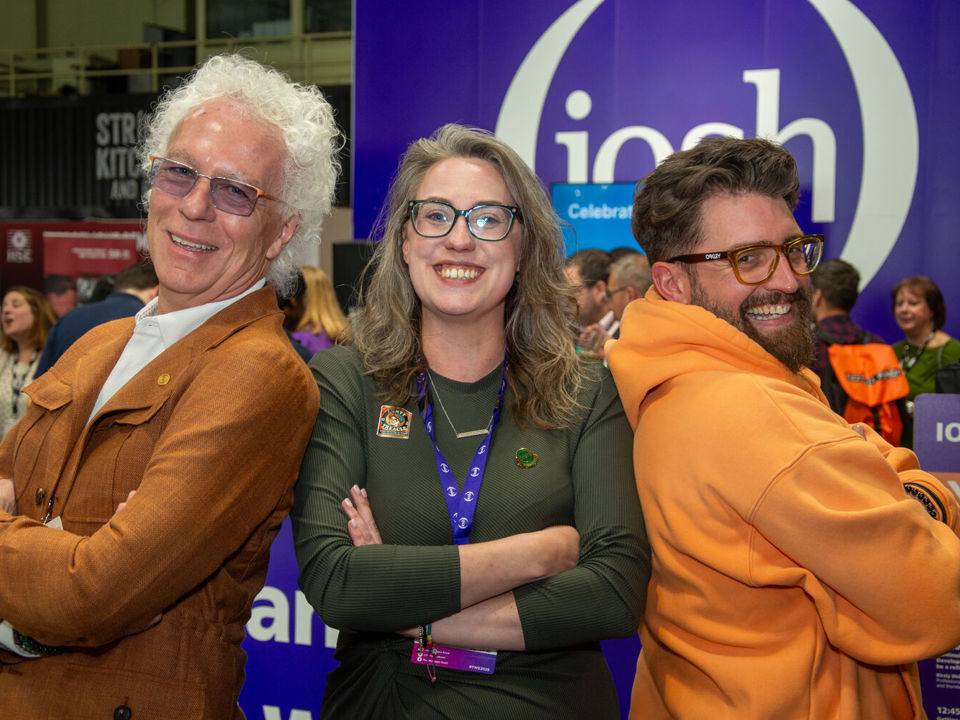 Three people standing back to back in front of an IOSH event display, posing confidently with their arms folded in a busy exhibition setting.