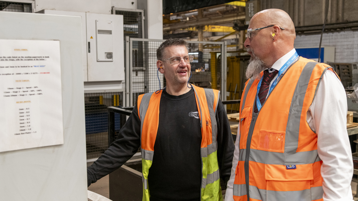Two employees wearing high-visibility vests and safety glasses discussing work beside industrial machinery on the Don-Bur factory floor, with safety information displayed on a nearby panel.