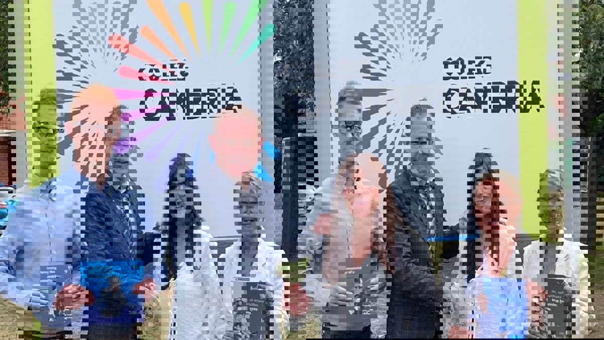 Four people standing outdoors in front of a large sign that reads “Coleg Cambria” with a colorful sunburst logo. Two individuals are holding booklets, and two are shaking hands. Trees and grass are visible in the background.