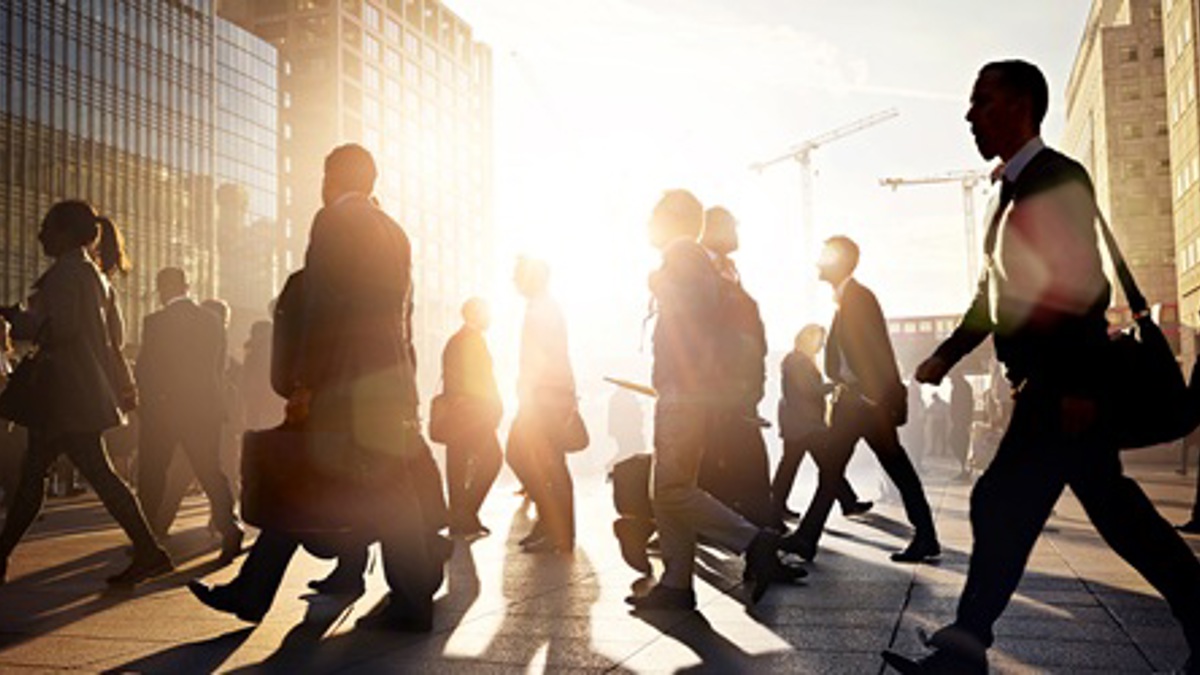 Crowd of people walking through a city street at sunrise, with tall modern buildings, long shadows on the ground and bright sunlight shining between the structures.