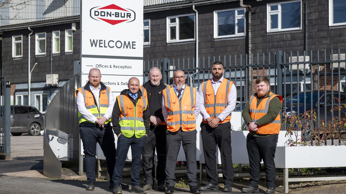 Group of Don-Bur employees wearing high-visibility vests standing outside the site entrance beside a large Don-Bur welcome sign.
