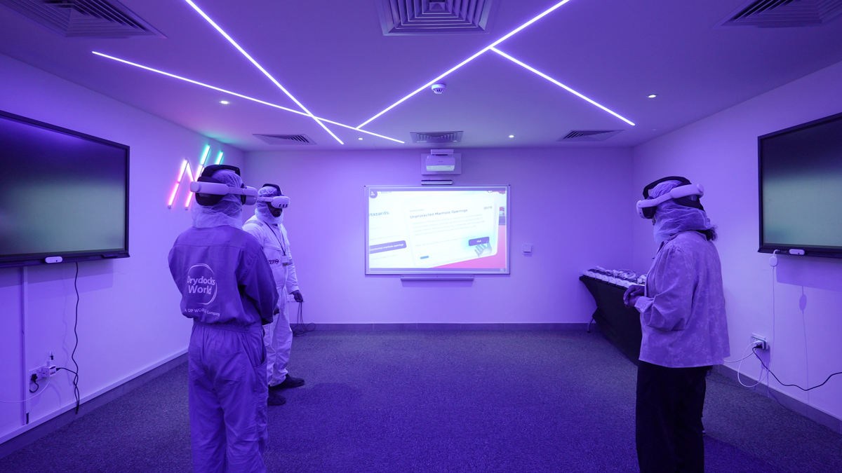 A VR training session taking place in a modern room illuminated with purple lighting and diagonal LED strips on the ceiling. Four individuals are wearing VR headsets and standing apart, engaged in the simulation. The room features two large wall-mounted screens on either side and a central projection screen displaying training content. A table with equipment is positioned along the right wall, and the floor is carpeted in gray.