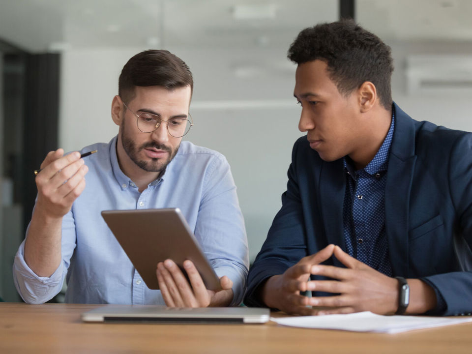 Two men sitting at desk in discussion and looking at tablet computer