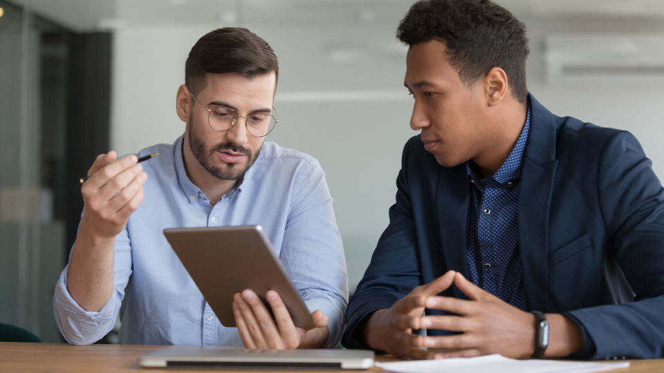 Two men sitting at desk in discussion and looking at tablet computer