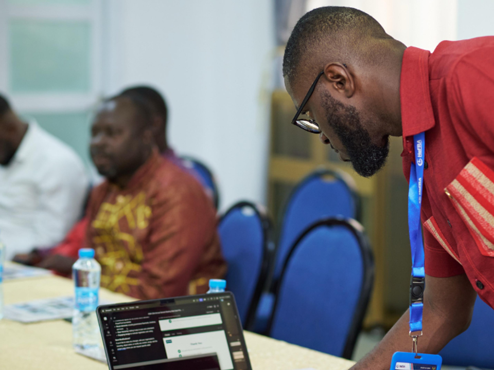A person wearing a red shirt and a lanyard leans over a table while using a laptop, with several other seated participants and blue chairs visible in the background. The scene takes place in a meeting or training room with documents and bottled water on the table, and the HeFRA logo displayed on the wall.
