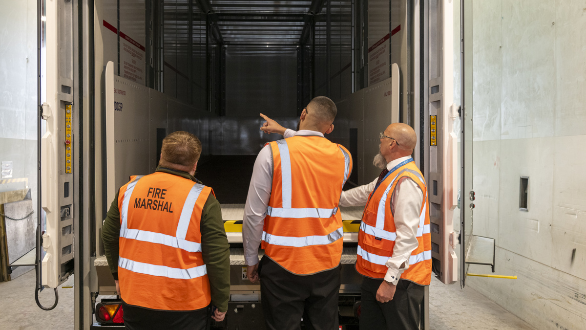 Three employees wearing high-visibility vests standing at the rear of a trailer inside a loading bay, inspecting the interior, with one pointing inside and a “Fire Marshal” label visible on one vest.