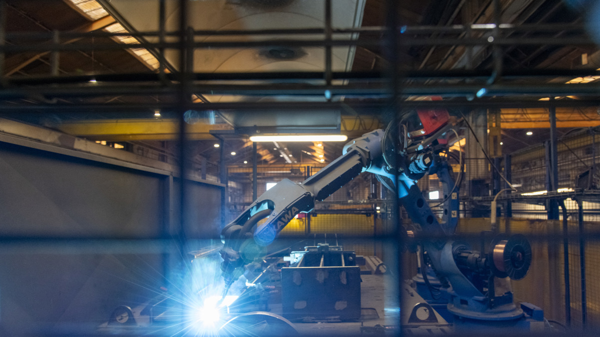 Industrial robotic arm welding metal components inside the Don-Bur factory, with bright sparks visible behind a protective safety barrier.